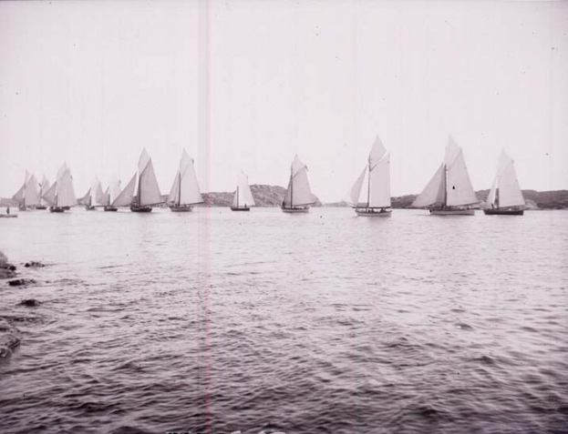 Schwarzes und weißes Foto von Segelbooten auf dem Wasser mit Hügeln im Hintergrund und einem klaren Himmel, als Foto des New York Yacht Club etikettiert.