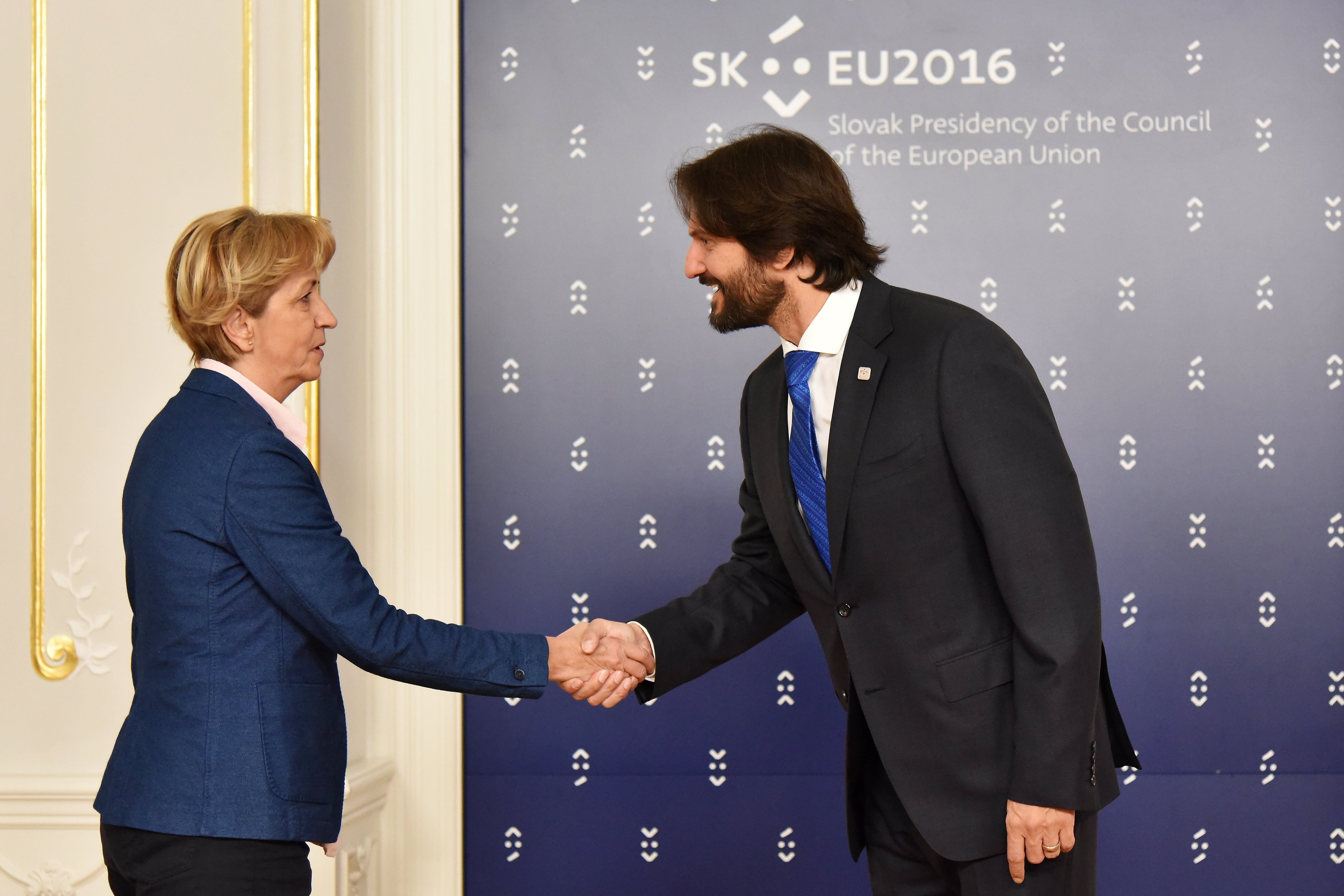 Bundeskanzlerin Angela Merkel und Bundespräsident Joachim Gauck schütteln sich die Hände und lächeln bei einem EU-Gipfeltreffen, mit einem "EU-Gipfeltreffen"-Banner und einer Tür im Hintergrund.