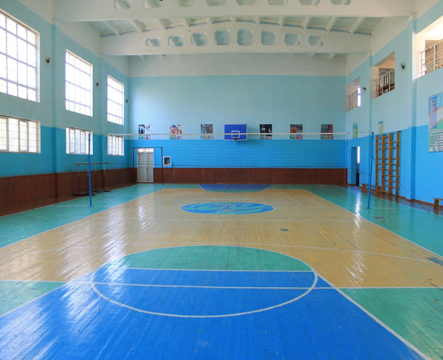 Eine große Turnhalle mit einem blauen und weißen Boden, die einen Basketballplatz, Netz, Stangen, eine Tür, Fenster, Plakate an der Wand, ein Geländer und ein Dach zeigt.