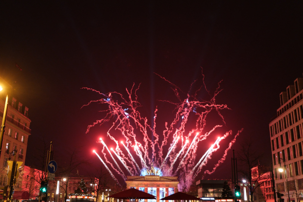Eine belebte Straßenkreuzung in Berlin an Silvester, voller Menschen, Fahrzeuge und festlicher Dekoration, mit Feuerwerk, das den Nachthimmel erhellt.