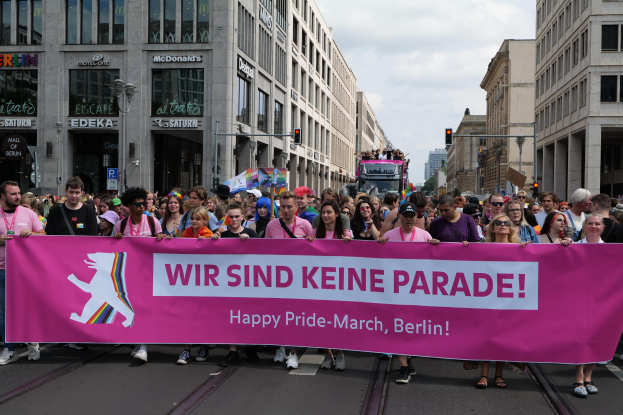 Eine Gruppe von Menschen marschiert auf einer Straße in Berlin mit einer pinken Fahne, auf der "Happy Pride March" steht, mit Gebäuden, Laternenmasten und Verkehrszeichen an der Straße.