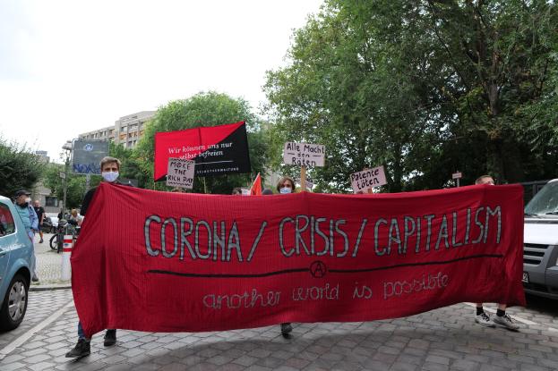 Eine Gruppe von Menschen marschiert mit einer roten Fahne, auf der 'Corona-Krise/Kapitalismus' steht, eine Straße entlang, mit parkenden Autos auf beiden Seiten und Gebäuden, Bäumen und einem klaren Himmel im Hintergrund.