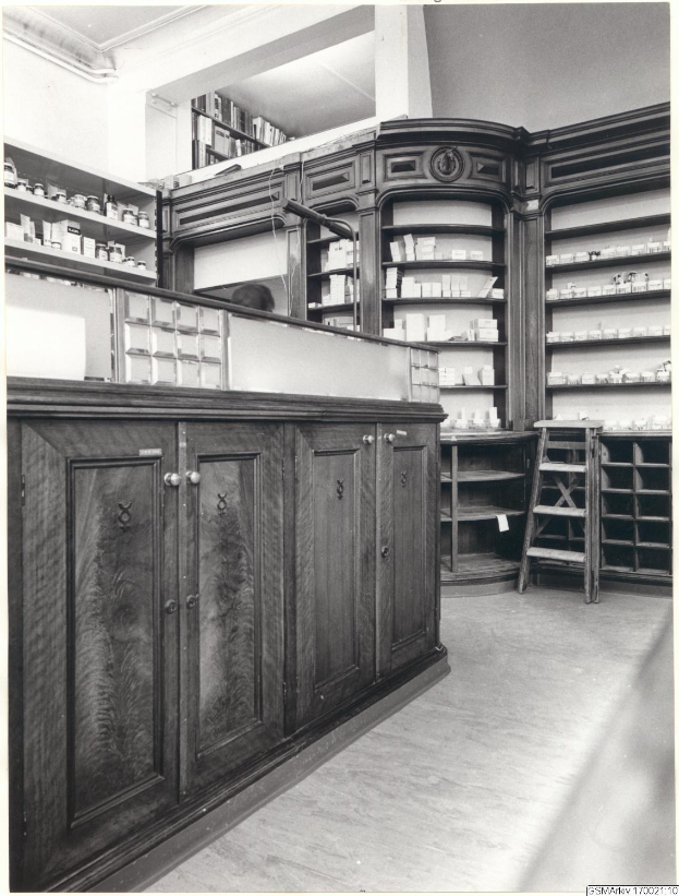 Schwarz-weiß-Foto eines vintage-Apotheken-Interieurs mit einer Theke, Schränken, einer Leiter und Regalen, die mit verschiedenen Gegenständen gefüllt sind, mit Text oben.
