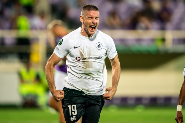 Ein Mann in weißem Hemd und schwarzen Shorts rennt auf einem Fußballfeld, umgeben von einer Gruppe von Menschen, die ein Tor feiern.