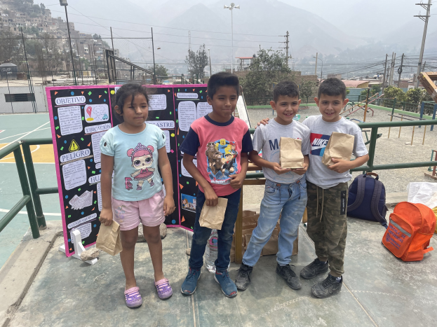 Gruppe von Kindern auf einem Basketballplatz mit Papiertüten, mit Tüten und Gegenst├Ąnden rechts und einer Tafel mit Text links, vor einem Hintergrund aus Zaun, Masten, Stra├člaternen, B├Ąumen, Geb├Ąuden, H├Ąngen und Himmel.