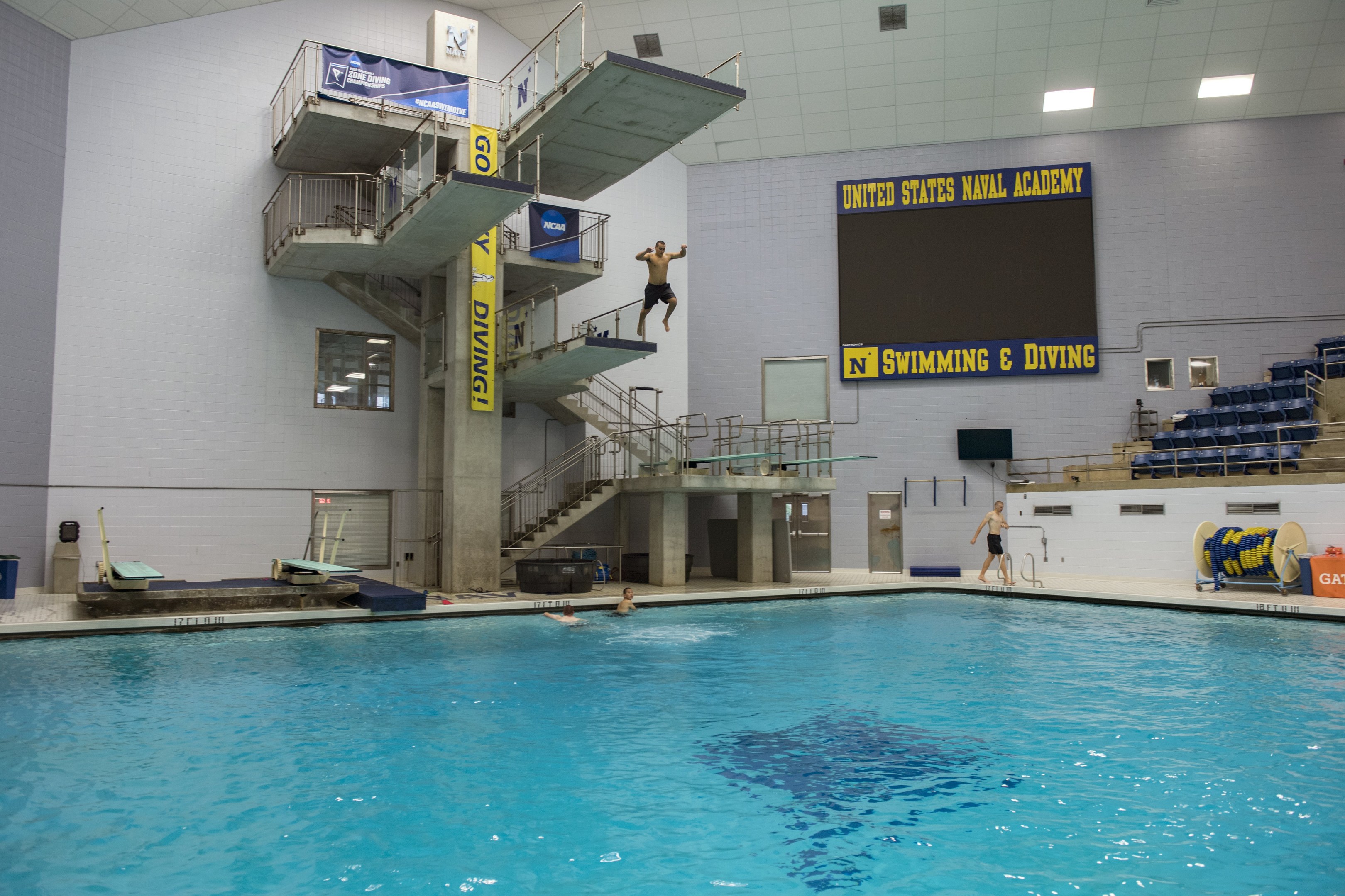 Großer Hallenbad mit schwimmenden Menschen, Treppe mit Geländer, Banner mit Text, eine Person springt ins Wasser, Stühle auf der rechten Seite, ein Bildschirm an der Wand und Deckenbeleuchtung.