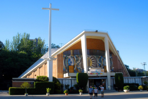 Eine Kirche mit einem großen Kreuz an der Front, umgeben von Topfpflanzen, einer Gruppe von Menschen, einem Metallzaun, Pfosten, einem Schild, einer Fahne mit Text, Gebäuden, Bäumen und einem bewölkten Himmel.