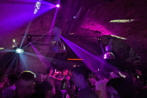 Eine Gruppe von Menschen, die in einem Nachtclub tanzen, beleuchtet von lila und blauen Deckenlichtern, einige halten Flaschen.