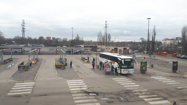 Ein Bus hält an einer Bushaltestelle mit Menschen drumherum, Fahrzeugen auf der Straße, Polen, Lampen, Schildern und anderen Gegenständen im Vordergrund, mit Bäumen, Gebäuden, Türmen mit Drähten und einem bewölkten Himmel im Hintergrund.
