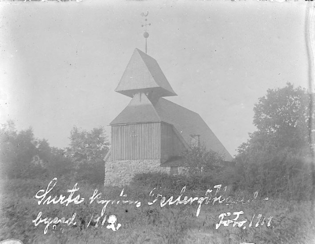 Schwarzes und weißes Foto der Kirche des Heiligen Grabes, eine Kirche mit einem Turm, umgeben von Bäumen und Pflanzen unter einem bewölkten Himmel.