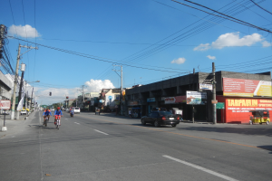 Eine belebte Stra├če mit Menschen auf Fahrr├Ądern und Autos, Strommasten mit Kabeln, Geb├Ąude mit Namensschildern und ein bew├Âlktes Himmel ├╝berall.