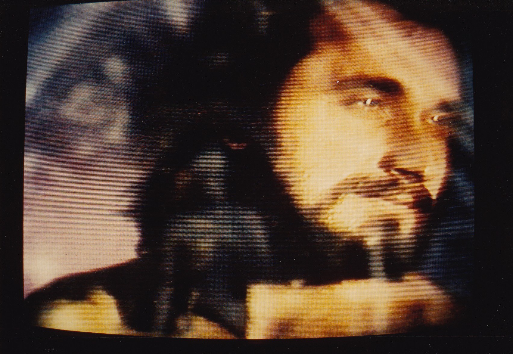 Ein Mann mit langem dunklem Bart und Schnurrbart, der ein weißes Hemd trägt, erscheint auf einem Fernseher mit dunklem Hintergrund und schaut direkt in die Kamera.