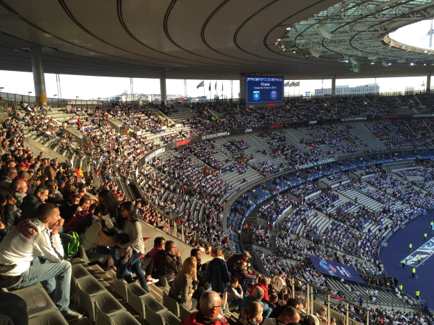Große Menschenmenge in einem Stadion bei einem Füssballspiel, mit einer Bühne auf der rechten Seite, Fahnen, Masten, einem Bildschirm und der Allianz Arena in München, Deutschland im Hintergrund.