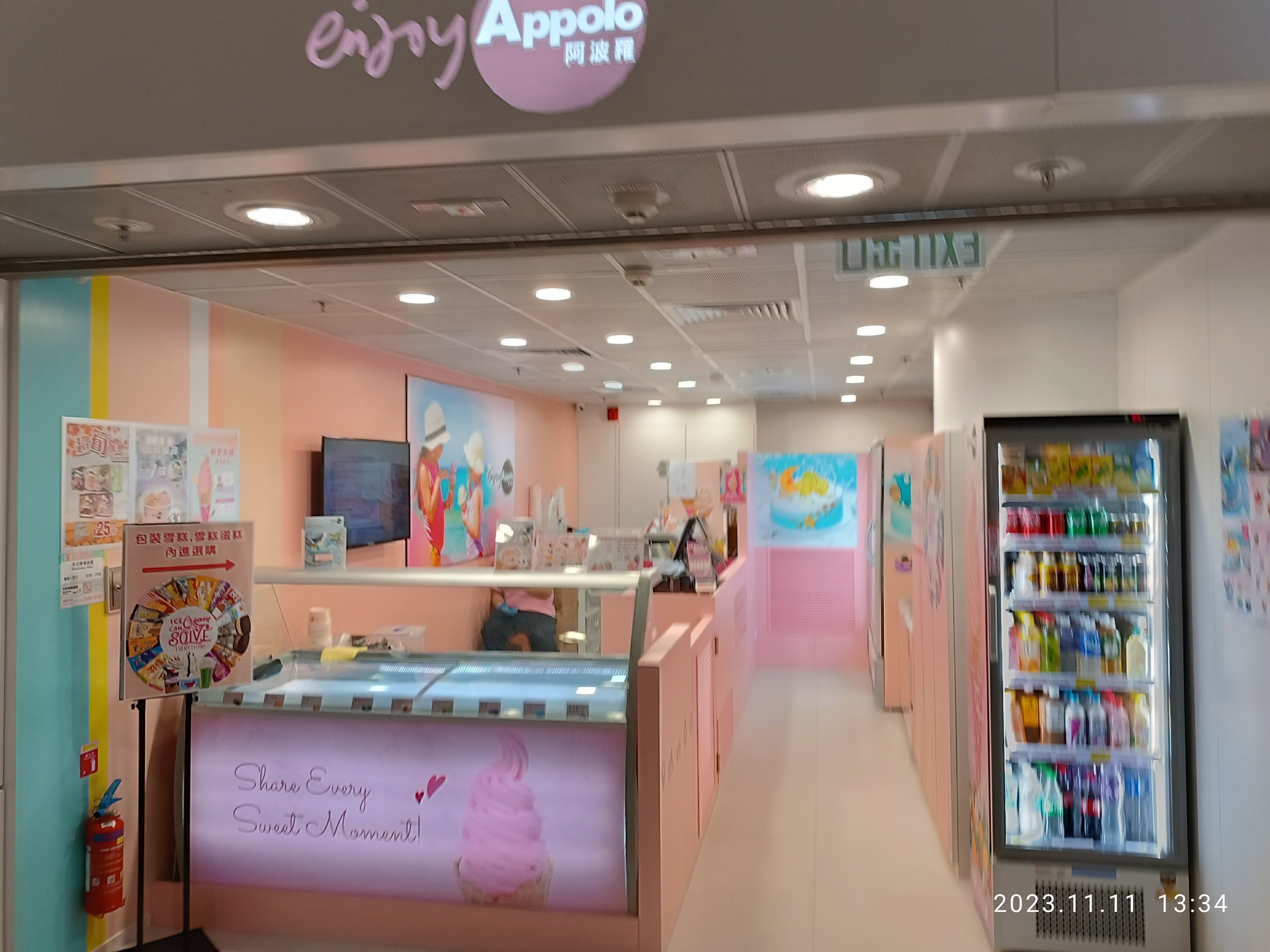 Innenansicht eines Eiscafés mit einem Kühlschrank auf der rechten Seite, der mit Flaschen gefüllt ist, einem Feuerlöscher und einer Tafel auf der linken Seite, einer Person an einem Tisch in der Mitte und einem Schild mit der Aufschrift 'Genießen Sie das Apollo-Eiscafé' an der Decke.