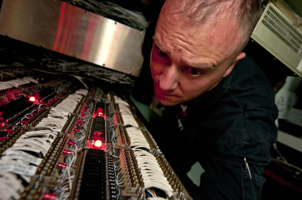 Ein Mann in einem schwarzen Hemd konzentriert sich auf einen Computerbildschirm, während er zwischen elektronischen Geräten und Kabeln arbeitet.