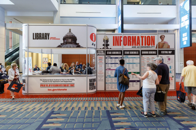 Menschen vor einem Stand auf dem Nationalen Buchfest, mit Bannern, Anzeigetafel und Deckenbeleuchtung.