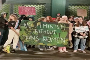 Gruppe von Menschen mit einem Banner, das "Kein Feminismus ohne Trans-Frauen" und Schilder in einem öffentlichen Raum mit einer Wand und einem Rollladen im Hintergrund zeigt.
