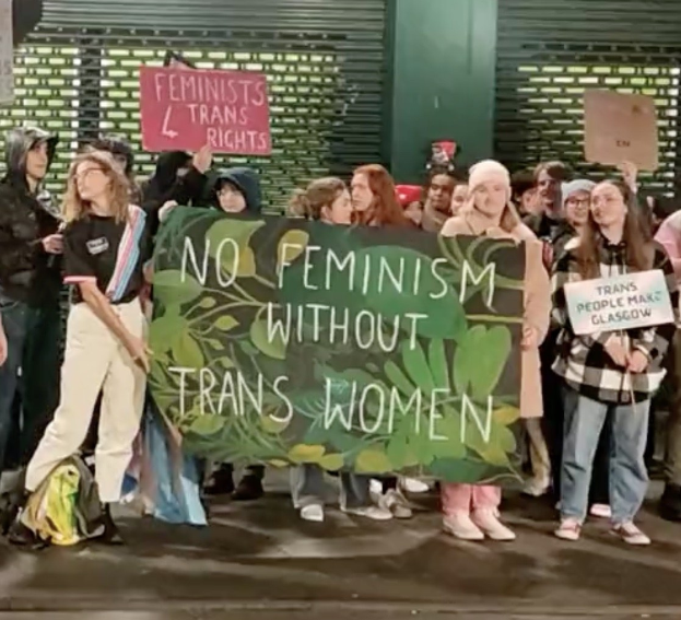 Gruppe von Menschen mit einem Banner, das "Kein Feminismus ohne Trans-Frauen" und Schilder in einem öffentlichen Raum mit einer Wand und einem Rollladen im Hintergrund zeigt.