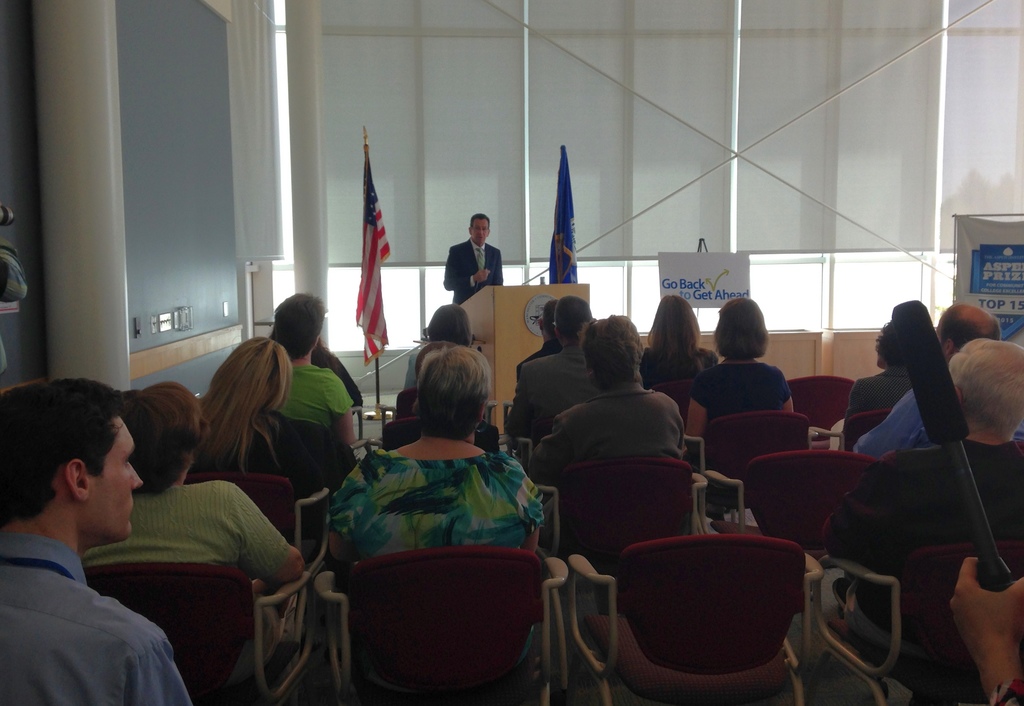 Ein Mann steht an einem Podium und spricht zu einem Publikum, das auf Stühlen sitzt, mit zwei Fahnen neben ihm und einer Wand, Säulen und Fenstern im Hintergrund.