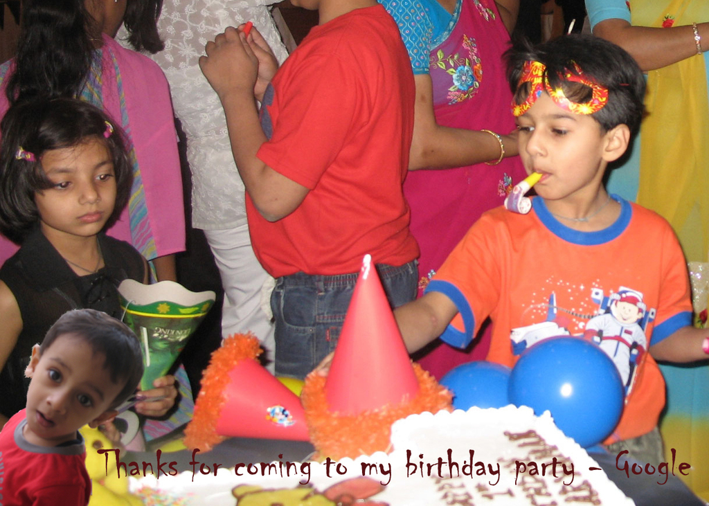 Kinder vor einem Tisch mit einem Kuchen, Luftballons und Mützen, mit anderen Menschen dahinter.