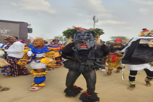 Eine Gruppe von Menschen in bunten Kostümen marschiert auf einer Straße bei der Lagos Carnival 2019, mit einer Person in einer Maske im Vordergrund und Bäumen, Pfählen und einem klaren blauen Himmel im Hintergrund.