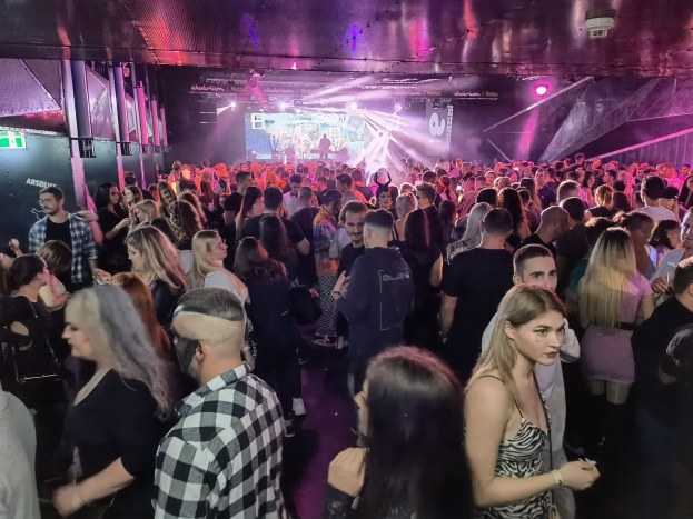Große Gruppe von Menschen in einer Diskothek mit Deckenbeleuchtung und Säulen, die an einem Absolut Nightlife-Event teilnehmen, mit einem Bildschirm im Hintergrund.