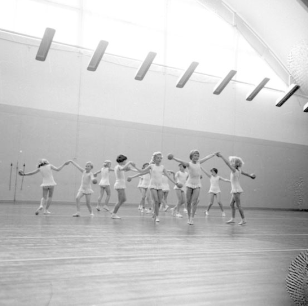 Schwarzes und weisses Bild von jungen Maedchen, die in einem Studio tanzen, sich an den Haenden halten und eine Kugel auf der rechten Seite und Lampen an der Decke haben.