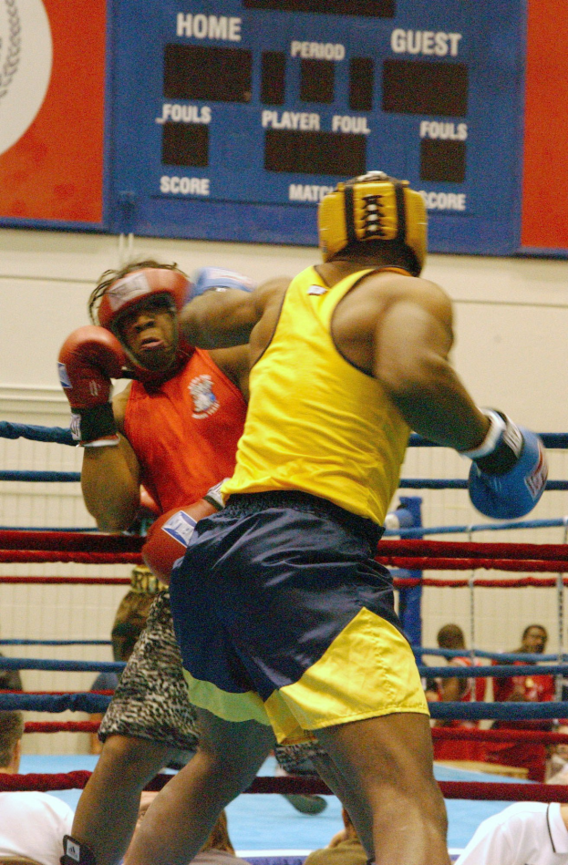 Zwei Boxer in einem Ring, die in einem Kampf gegeneinander antreten, mit Zuschauern im Hintergrund und einer Tafel mit Text und Logos an der Wand.