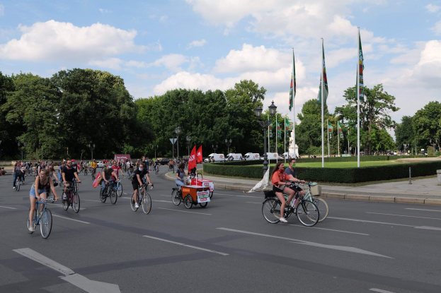 Eine Gruppe von Menschen, die auf Fahrrädern eine von Bäumen gesäumte Straße mit Laternenmasten und Fahnenmasten entlangfahren, während im Hintergrund Fahrzeuge und bewölkter Himmel zu sehen sind.