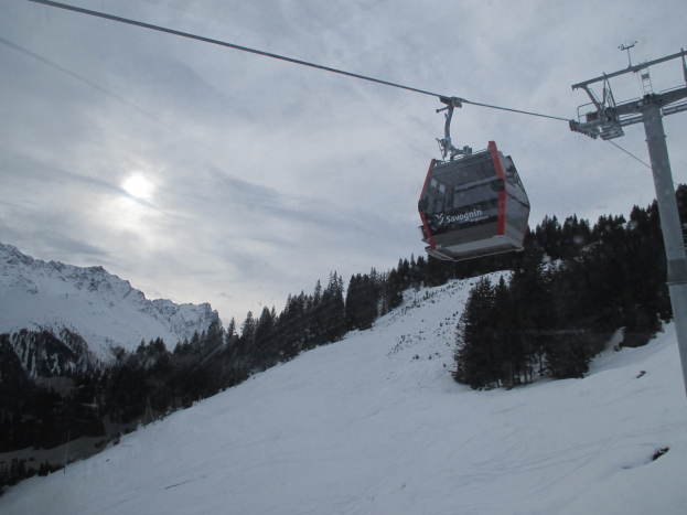 Eine Seilbahn fährt einen schneebedeckten Berg hinauf, mit Bäumen im Hintergrund, unter einem bewölkten Himmel mit hellen Sonnenstrahlen.