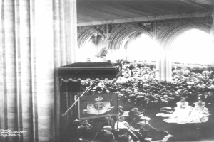 Ein Schwarz-Wei├č-Foto einer gro├čen Menge in einer Kirche, mit einem Podium, das mit einer Flagge drapiert und von Lichtern beleuchtet ist.