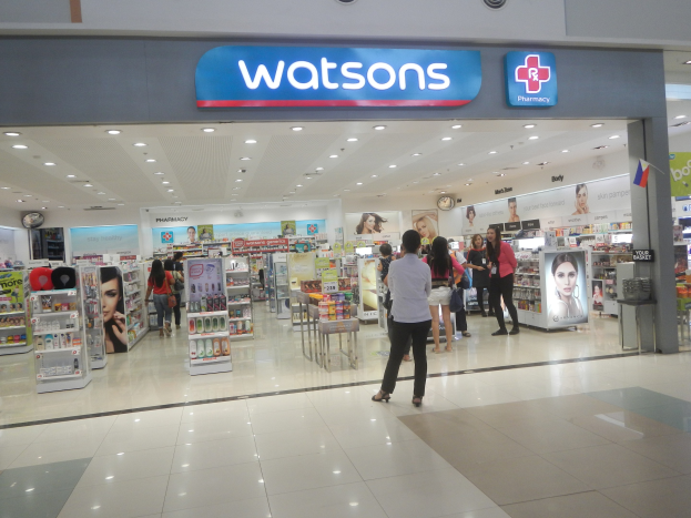 Eine Watsons Apotheke in einem Einkaufszentrum mit Menschen, Produkten, Regalen, Schildern, einer Wanduhr und Deckenleuchten.