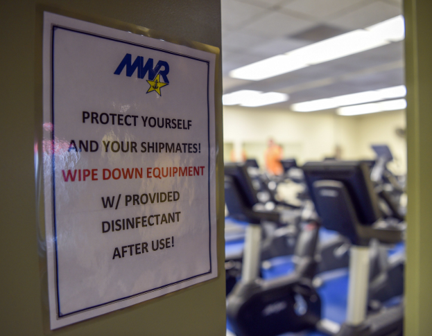 Ein Fitnessstudio-Türschild mit der Aufschrift "Schützen Sie sich und Ihre Schiffskameraden" mit unscharfen Laufbändern und Deckenleuchten im Hintergrund.