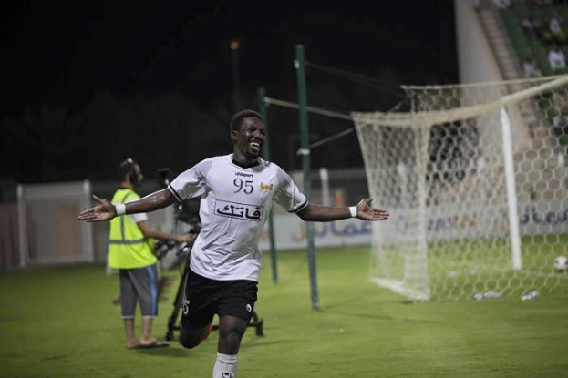 Ein Mann in weißem Hemd und schwarzen Shorts rennt über ein Fußballfeld, die Arme ausgestreckt, umgeben von Zuschauern, mit einem Torpfosten und einer sitzenden Menge im Hintergrund.