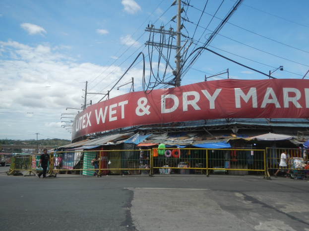 Nasse und trockene Marktstände entlang einer Straße mit einer Straße unten, ein paar Menschen auf der linken Seite, Pfähle und Drähte sichtbar und ein bewölkter Himmel.