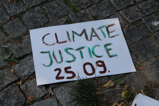 Weißes Schild mit schwarzer Schrift "Klimagerechtigkeit" auf Gras mit grünen und braunen trockenen Blättern.
