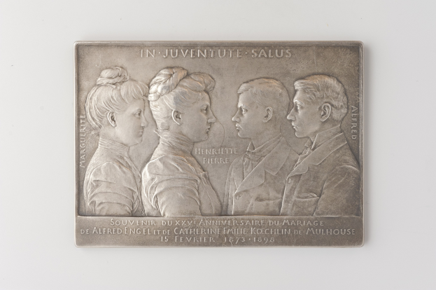 Silberne Plakette mit einem Schwarz-Weiß-Foto von drei Personen, graviert mit Text, vor einem weißen Hintergrund, wahrscheinlich zur Erinnerung an eine Hochzeit.