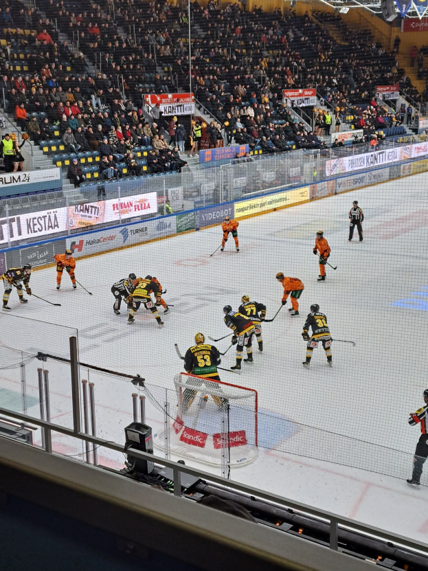 Eine Gruppe von Menschen, die Hockey auf einer Indoor-Eisbahn spielen, umgeben von einem Zaun, mit Zuschauern im Hintergrund und beleuchteter Stadionbeleuchtung über ihnen.