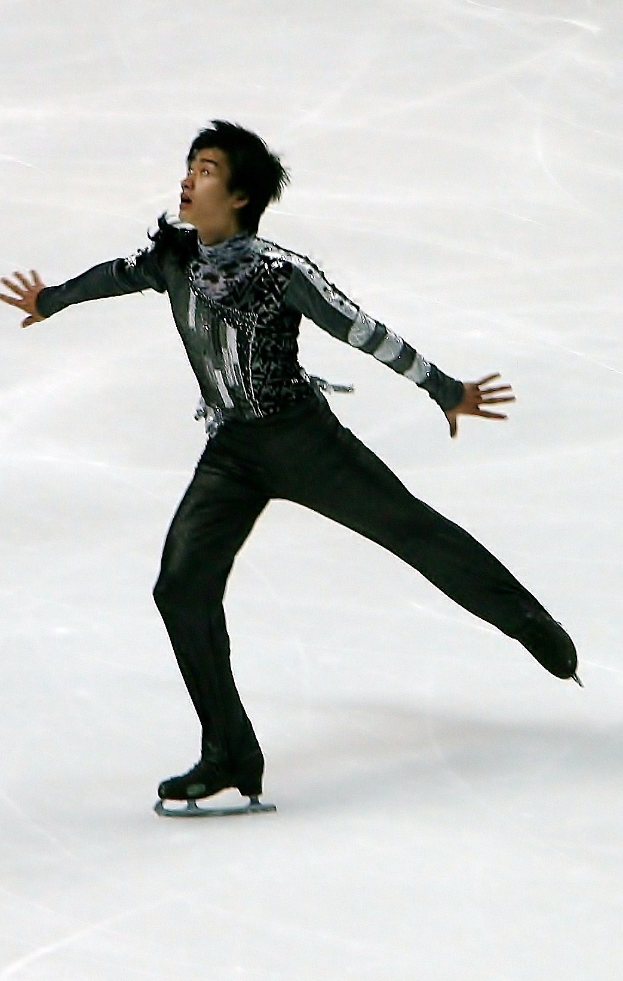 Männlicher Eiskunstläufer in schwarzer Kleidung gleitet über eine Eisfläche und führt eine Choreografie mit synchronen Arm- und Beinbewegungen aus.