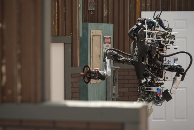 Ein humanoider Roboter mit ausgestreckten Armen und geneigtem Kopf steht vor einem Gebäude, seine großen runden Augen und der leicht geöffnete Mund deuten darauf hin, dass er sprechen könnte.