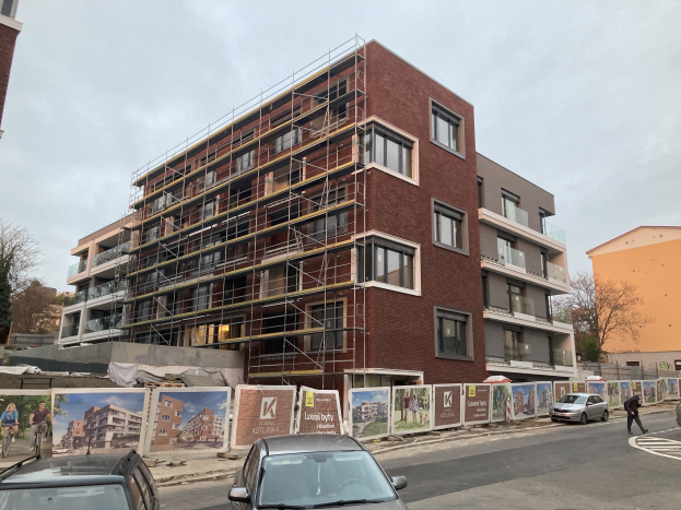 Großes Gebäude mit Gerüst an der Seite, umgeben von Bäumen unter einem klaren blauen Himmel, mit Fahrzeugen auf der Straße und einer Person auf dem Gehweg.