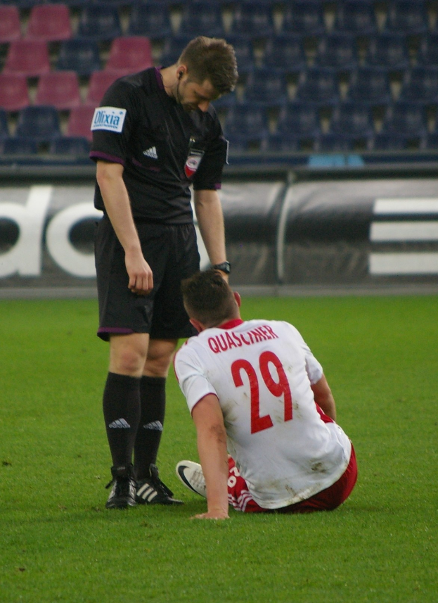 Ein Fussballspieler In Sportbekleidung Sitzt Auf Dem Boden Neben Einem Schiedsrichter In Einer Stadionumgebung.