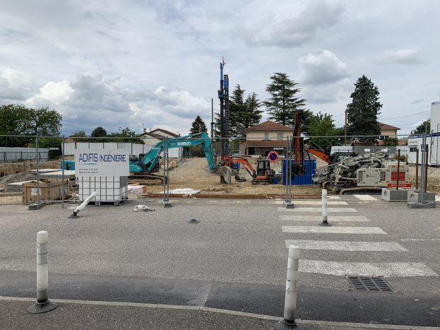 Baustelle mit verschiedenen Geräten, einschließlich Pfosten, einem Zaun, einem Schild, Fahrzeugen, Bäumen, Häusern und einem bewölkten Himmel.
