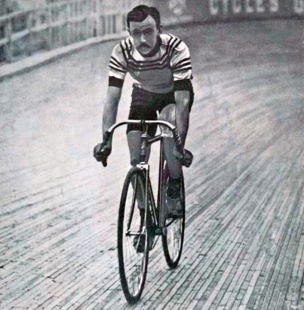 Schwarz-weiß-Foto eines Mannes, der auf einer Bahn radfährt, mit einem Zaun und einer Tafel im Hintergrund.