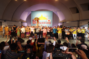 Eine Gruppe von Menschen auf einer Bühne mit Kameras, Handys und Musikinstrumenten während der Eröffnungszeremonie eines Festivals, mit einem Banner, Wänden und Deckenlampen im Hintergrund.