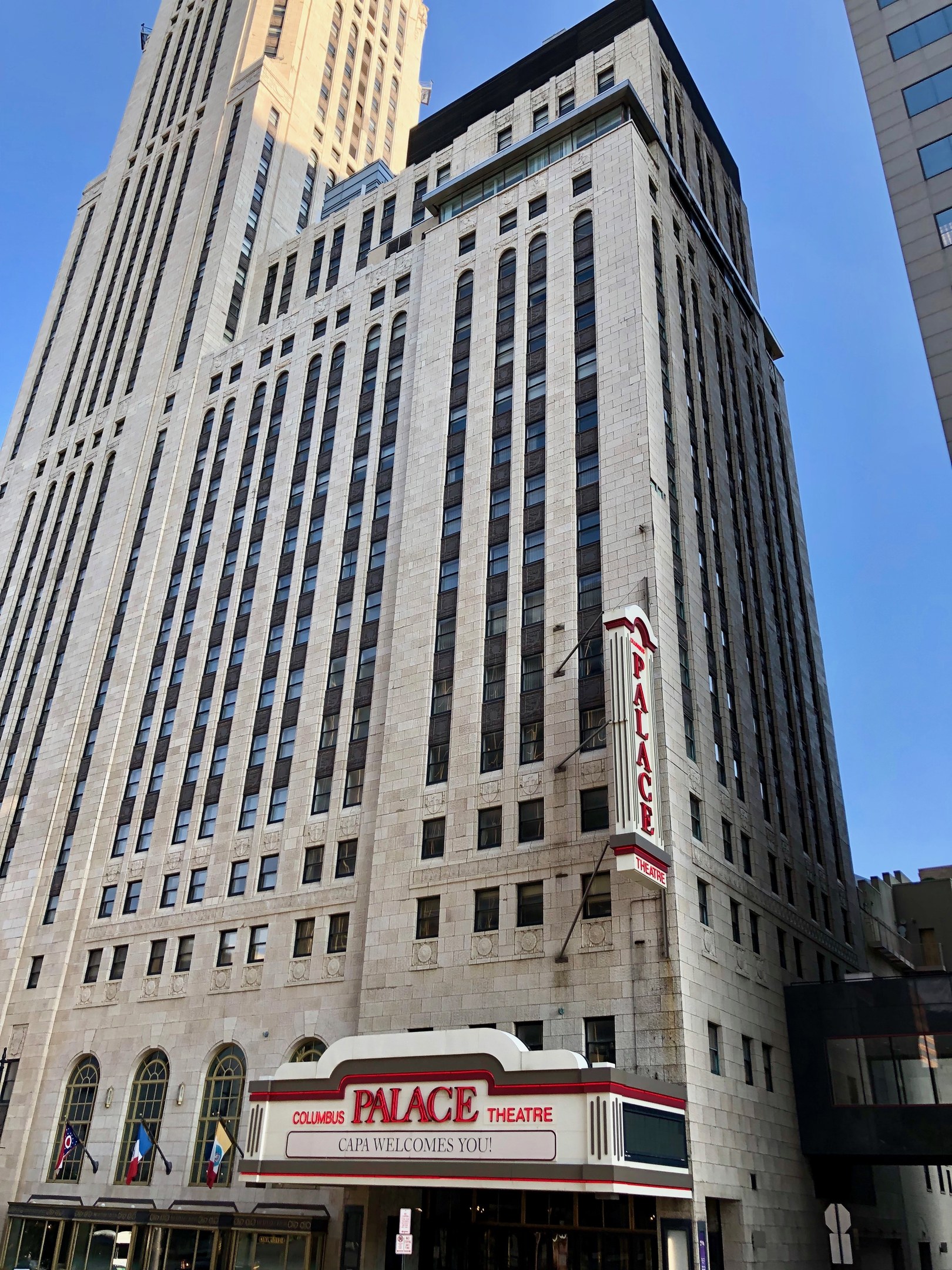 Außenansicht des Palace Theatre in New York City mit hohen Gebäuden, Fenstern, Namensschildern, Flaggen, Schildern, Fahrzeugen und Himmel im Hintergrund.