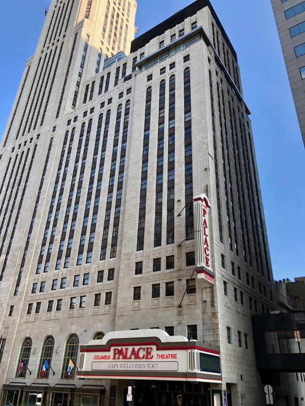 Außenansicht des Palace Theatre in New York City mit hohen Gebäuden, Fenstern, Namensschildern, Flaggen, Schildern, Fahrzeugen und Himmel im Hintergrund.