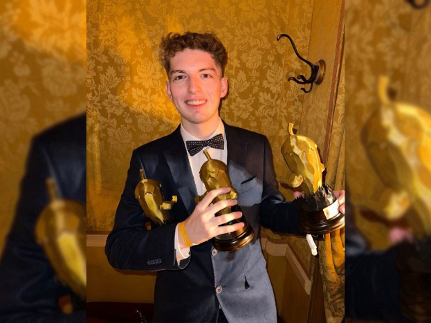 Ein Mann im Smoking lächelt und hält eine Oscar-Statue in der Hand, mit einer Wand im Hintergrund und einem Wasserzeichen auf dem Bild.