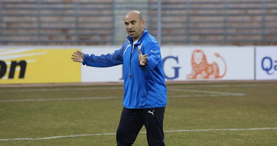 Ein Mann in einer blauen Jacke steht auf einem Fußballfeld während eines Trainings, umgeben von Tafeln mit Text und Logos, entschlossen wirkend.