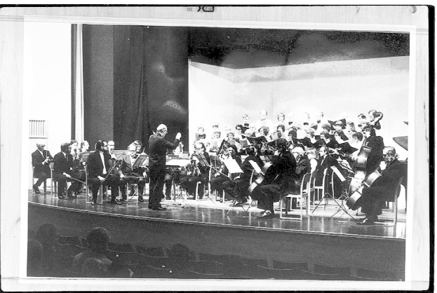 Ein Dirigent in Anzug und Krawatte steht auf einer Bühne und leitet ein Orchester mit Geigen, Celli und Flöten, während das Publikum auf Stühlen sitzt und aufmerksam zuhört.